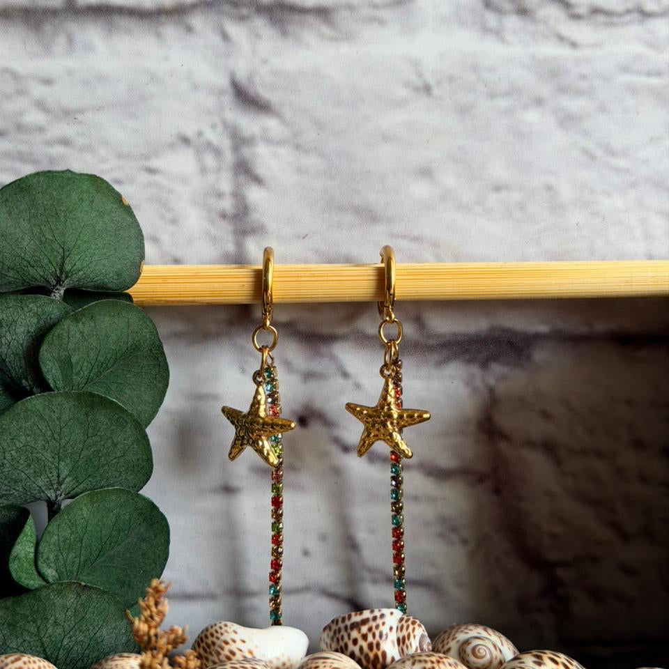 Gold starfish earrings with colorful beads hanging on a wooden stick, surrounded by shells and greenery.